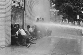 Black and white photo of students getting assaulted with water from a fire hole/ 